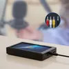 A portable video recorder with a screen and cables, placed on a table in front of a blurred background of a person and a microphone. Náhled obrázku produktu