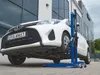 A white car is being lifted by a blue hydraulic jack in front of a modern building. Produktbild-Vorschau