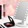 A collection of makeup products including a lighted makeup mirror, mascara, lipstick, and blush, arranged on a white surface with a soft pink, bokeh background. Voir les images du produit