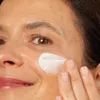 A close-up of a woman's face as she applies cream to her cheek. Podgląd zdjęcia produktu