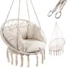 A macrame hanging chair with a cushion and tassels, shown on a white background. A smaller, empty macrame chair is shown in the background, along with metal rings and rope. Podgląd zdjęcia produktu