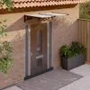 A door canopy is installed above a front door, with a planter and plants to the side. Náhľad obrázku produktu