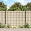 A beige PVC garden privacy screen with a grid pattern, set against a backdrop of green trees and a blue sky. Plants and a paved walkway are visible in the foreground. Voir les images du produit