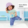 A doll is standing on one leg, wearing a purple hat, green checkered shirt, and blue jeans. Produktbild-Vorschau