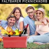 Four people are having a picnic in a park, with a picnic basket in the foreground. Produktbild-Vorschau