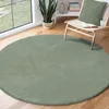A round, green rug is placed on a wooden floor in a room with a large window and a chair. Produktbild-Vorschau
