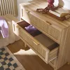 A close-up view of a wooden dresser with rattan drawer fronts, showing two open drawers with clothing and a box inside. A decorative tray with books and a pink vase sits on top. Produktbild-Vorschau