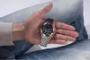 A person's hand holding a watch, with jeans in the background. Náhled obrázku produktu