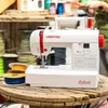 A white sewing machine with red accents sits on a wooden spool, surrounded by spools of thread and ribbon. Produktbild-Vorschau