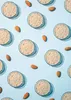 A repeating pattern of small glass containers filled with almond flour and whole almonds scattered on a light blue background. Produktbild-Vorschau