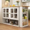 A white kitchen island with glass-fronted cabinets and open shelving is presented in a well-lit room. Produktbild-Vorschau