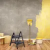 A room being renovated with a wall partially painted yellow, a ladder, paint cans, and boxes. Voir les images du produit