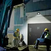 Two construction workers in a dimly lit industrial area at night, with one worker operating a piece of machinery and the other standing nearby. Produktbild-Vorschau