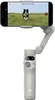 A smartphone is mounted on a white gimbal stabilizer, with a dog running in a field visible on the phone screen. Voir les images du produit