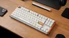 A mechanical keyboard is placed on a wooden desk with a smartphone and headphones nearby. Produktbild-Vorschau