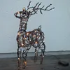 A lighted reindeer decoration made of wire with a rattan look, placed on a wooden surface against a textured wall. Podgląd zdjęcia produktu