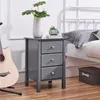 A gray nightstand with three drawers is placed next to a bed. A plant in a white vase and a green decorative object are on top of the nightstand. A guitar is leaning against the nightstand. Produktbild-Vorschau