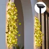 A solar-powered string of lights with artificial ivy leaves is draped along a white wall, with a close-up inset of the solar panel. Podgląd zdjęcia produktu
