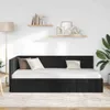 A black velvet daybed with white bedding and pillows, set against a white wall with two wooden shelves displaying decor and books. A floor lamp is visible to the left, and a large plant to the right. Produktbild-Vorschau