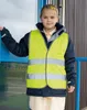 A young girl wearing a safety vest over a jacket and school uniform stands by a building entrance. Produktbild-Vorschau