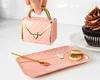 A hand holding a pink handbag-shaped mug over a matching pink tray with cookies and a spoon, with a cupcake in the background. Produktbild-Vorschau