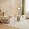 A white wooden children's bed with a fence and a door, set up in a room with decorative pictures on the wall and a floor lamp. Náhľad obrázku produktu