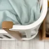 A close-up view of the edge of a white baby crib with a wooden accent and a light blue fabric draped inside. Produktbild-Vorschau
