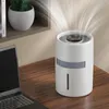 A white air purifier/humidifier is shown on a wooden desk next to a laptop, with a plant in a pot on a shelf in the background. White wisps of air are shown emanating from the top of the device. Náhled obrázku produktu
