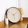 A minimalist wall clock with a beige face and black frame is displayed on a wooden surface next to a small vase with dried flowers and a pleated lamp. Anteprima dell’immagine prodotto