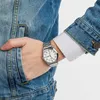 A person wearing a denim jacket and a white shirt, showing off a silver watch on their wrist. Produktbild-Vorschau