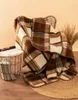A plaid wool blanket draped over a chair, with a magazine rack and dried grass in the background. Produktbild-Vorschau