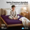 A young boy sitting cross-legged on a purple bedsheet, with pillows behind him and a blanket draped over the side of the bed. A bedside table with a lamp and mug is visible. Produktbild-Vorschau