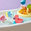 A bathtub with several floating toys, including a flamingo, a fish, an octopus, and a lion on a yellow platform. Water is being poured from a blue cup onto the lion. Náhled obrázku produktu
