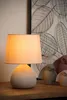 A close-up shot of a beige table lamp with a textured base and a light-colored lampshade. Behind the lamp, there are decorative owl figurines and a wall art with leaf patterns. Náhled obrázku produktu