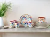 A collection of ceramic tableware with a colorful floral pattern, including plates, bowls, and mugs, arranged on a wooden shelf. Náhľad obrázku produktu