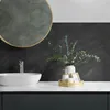 A bathroom vanity with a sink, faucet, and decorative items on the countertop, with a dark herringbone tile backsplash and a round mirror above. Produktbild-Vorschau