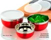 Three red stainless steel bowls with white lids, one filled with green leaves, on a wooden cutting board. Produktbild-Vorschau