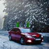 A red car decorated with Christmas trees on the roof is driving on a snowy road surrounded by snow-covered trees. Podgląd zdjęcia produktu
