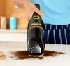 A person is using a black Kärcher handheld vacuum cleaner to clean up spilled coffee grounds on a white countertop. Produktbild-Vorschau
