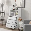 A white wooden dresser with three open drawers, a shelf with a speaker and decor, and a floor lamp in the background. Náhled obrázku produktu