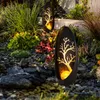 Two solar-powered garden lights with a tree silhouette design are placed in a garden setting with rocks and plants. Produktbild-Vorschau