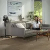 A woman sits on a couch in a living room with a fan in the foreground. Produktbild-Vorschau
