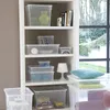 A white bookshelf filled with various clear storage boxes, some containing items like vases, books, and board games. A window with greenery is visible in the background. Produktbild-Vorschau