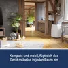 A white portable air conditioner is placed in a living room with wooden floors, a plush rug, and decorative items. Produktbild-Vorschau