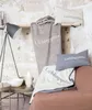A collection of home decor items, including blankets and pillows, arranged on a shelf and a sofa, with a lamp and side table in the foreground. Produktbild-Vorschau