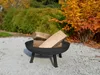 A black metal fire pit filled with logs sits on a gravel surface in a garden setting with autumn foliage in the background. Anteprima dell’immagine prodotto