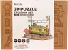 The image shows the packaging of a 3D puzzle creation set, featuring a model of a train station with a green train and cherry blossoms. Produktbild-Vorschau