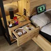A bedside table with a lamp, phone, headphones, and books, next to a bed. Produktbild-Vorschau