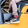 A person wearing Reebok work sneakers is sitting in the driver's seat of a construction vehicle. Náhľad obrázku produktu