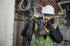 A construction worker is using a rotary hammer drill to drill into a concrete wall. The background shows exposed pipes and wiring, typical of a construction site. Produktbild-Vorschau